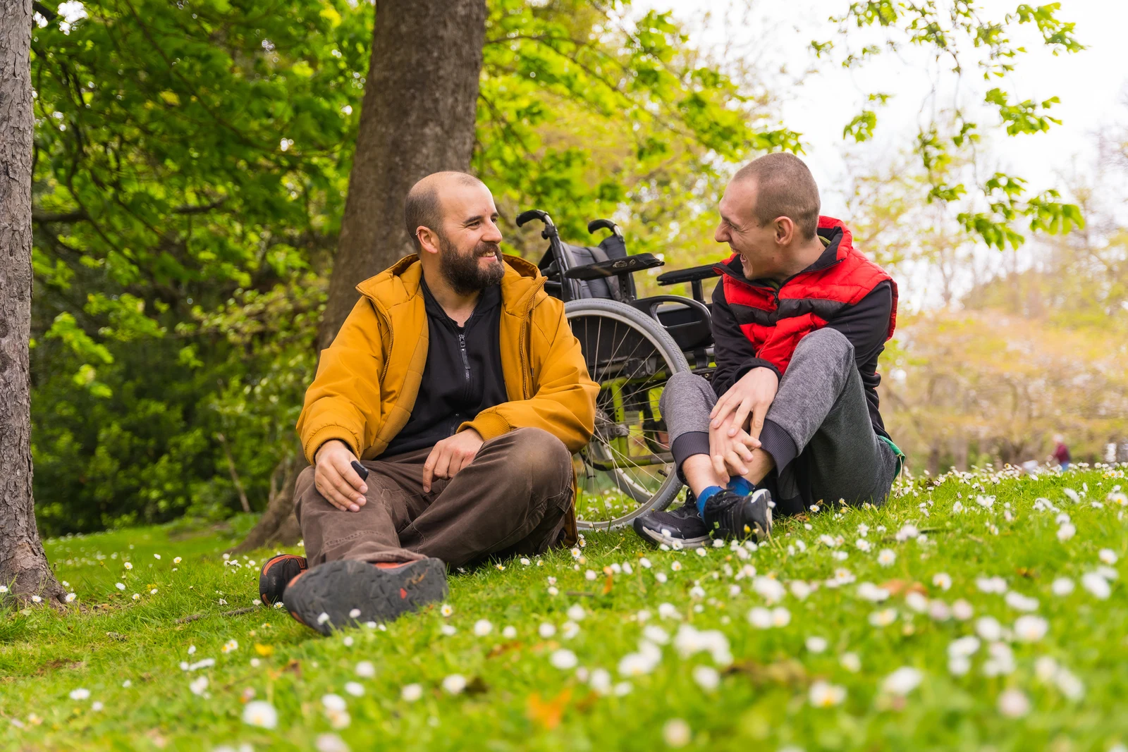 A paralyzed young man with a friend sitting on the grass of a public park in the city, talking and laughing